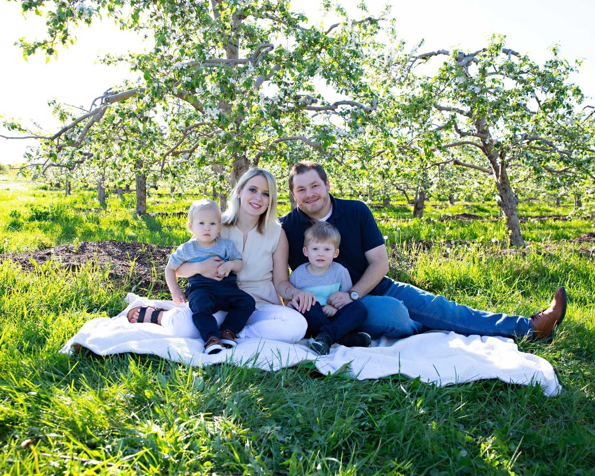 Family sitting on a blanket in a lush green orchard, smiling and enjoying the day.